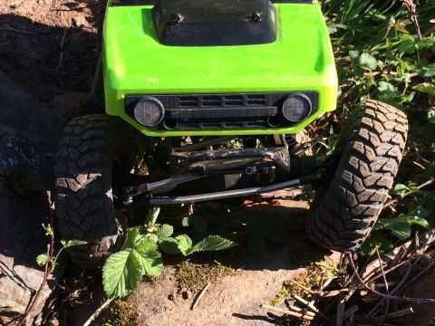 1989 Ford Bronco - A bright green RC rock crawler is shown from the front, navigating an outdoor terrain with rocks and plants.