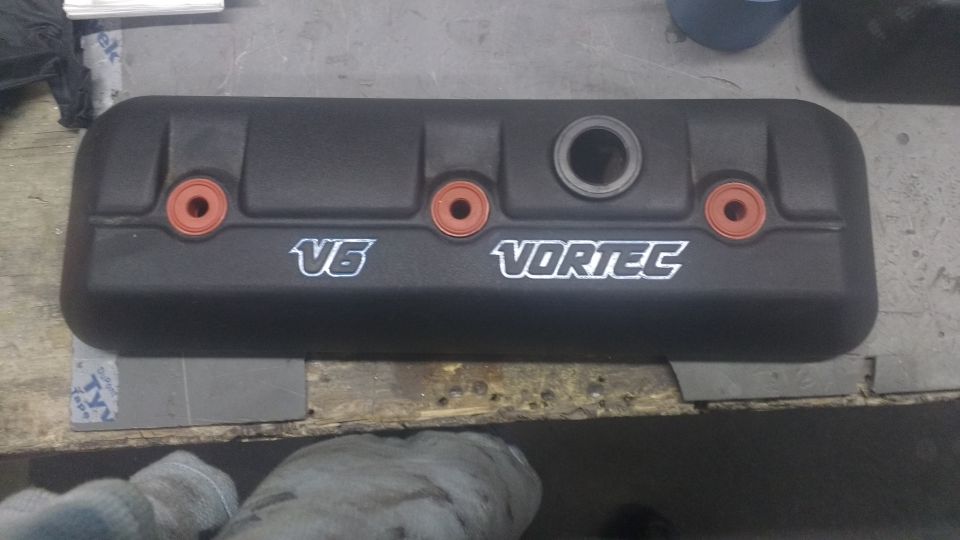 Black engine valve cover with V6 Vortec branding and orange grommets