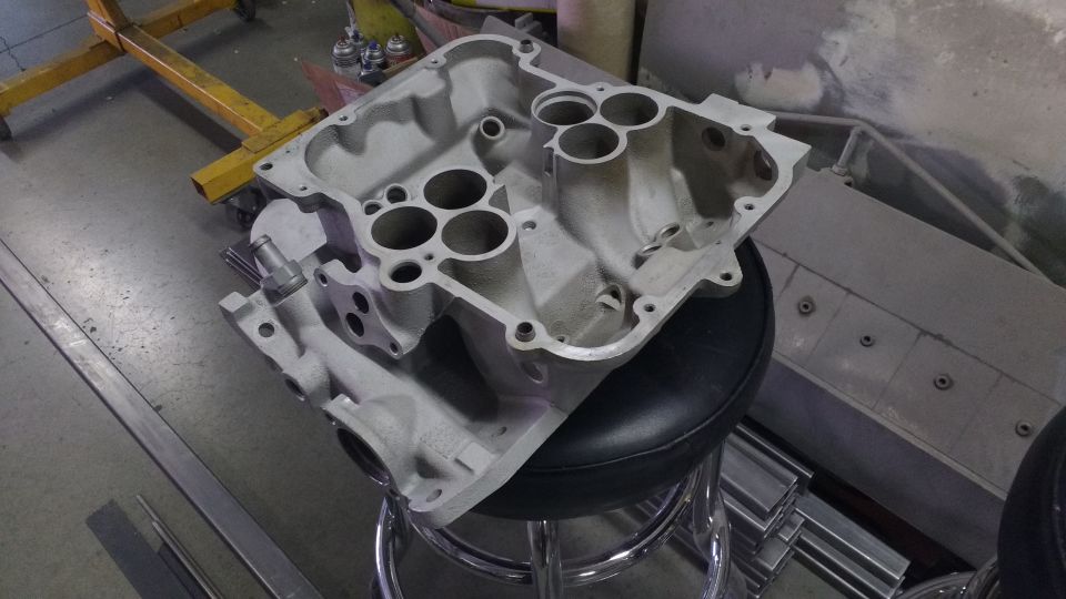Aluminum intake manifold on a stool, with a yellow engine hoist in the background