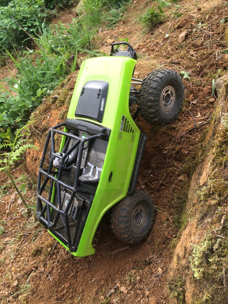 Green RC rock crawler tipped on its side on a dirt hill, with a roll cage visible