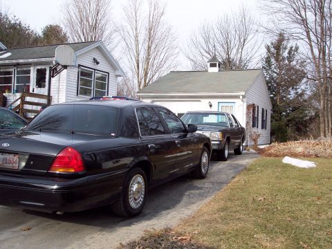 2001 Ford Crown Victoria - Two sedans are parked in a driveway in front of houses.
