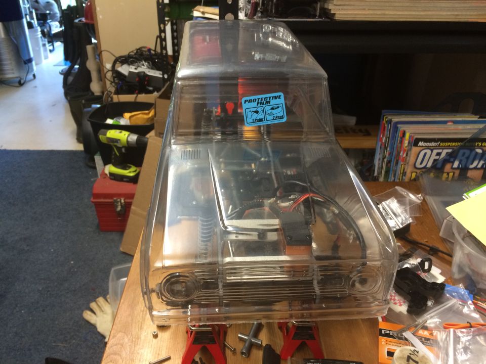 Clear plastic RC car body on red jack stands, showing internal components. Protective film sticker on windshield