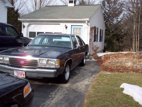 2001 Ford Crown Victoria - A dark sedan is parked in a driveway next to a garage, with another vehicle partially visible in the foreground.