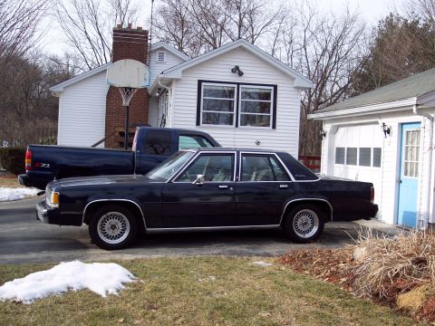 2001 Ford Crown Victoria - A dark sedan and a blue pickup truck are parked in a driveway next to a house.
