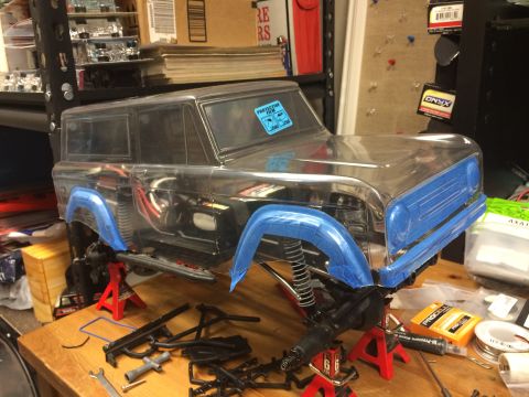 1989 Ford Bronco - Unassembled RC car body with protective film on the windshield sits on jack stands amidst tools and parts.