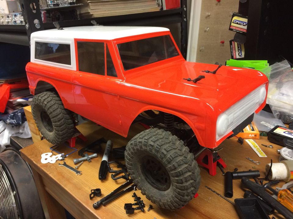 Orange and white RC car body resembling a Ford Bronco, on red jack stands with tools on workbench