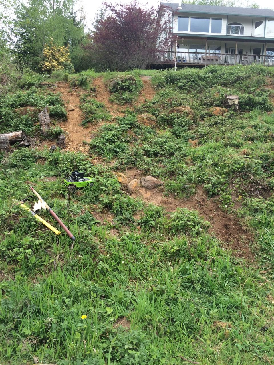 Green RC off-road vehicle on a grassy, muddy slope near some rocks and tools