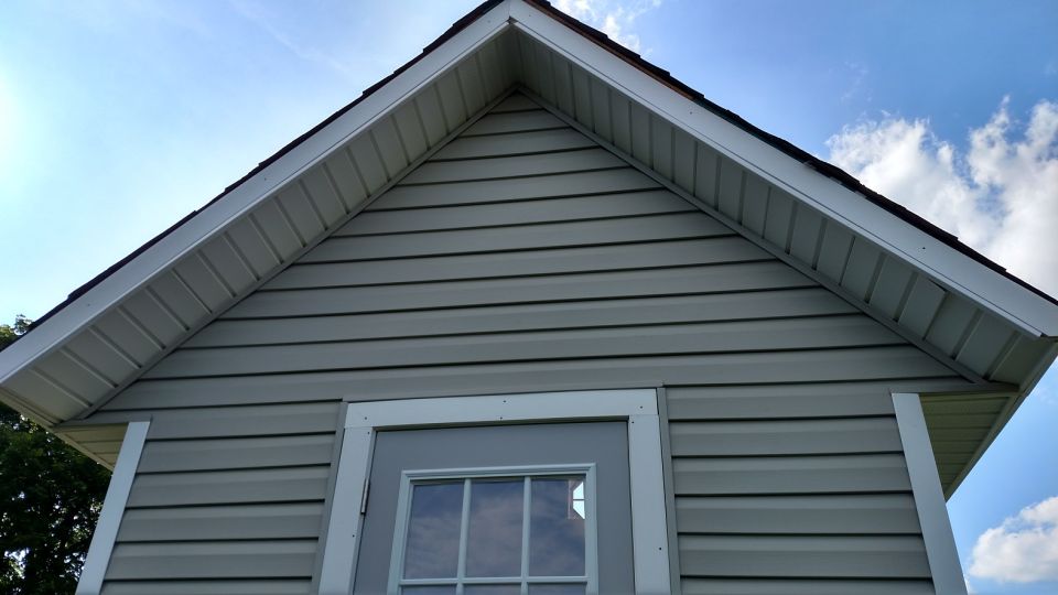 Exterior of a light gray playhouse with a white door and window, under a blue sky with clouds