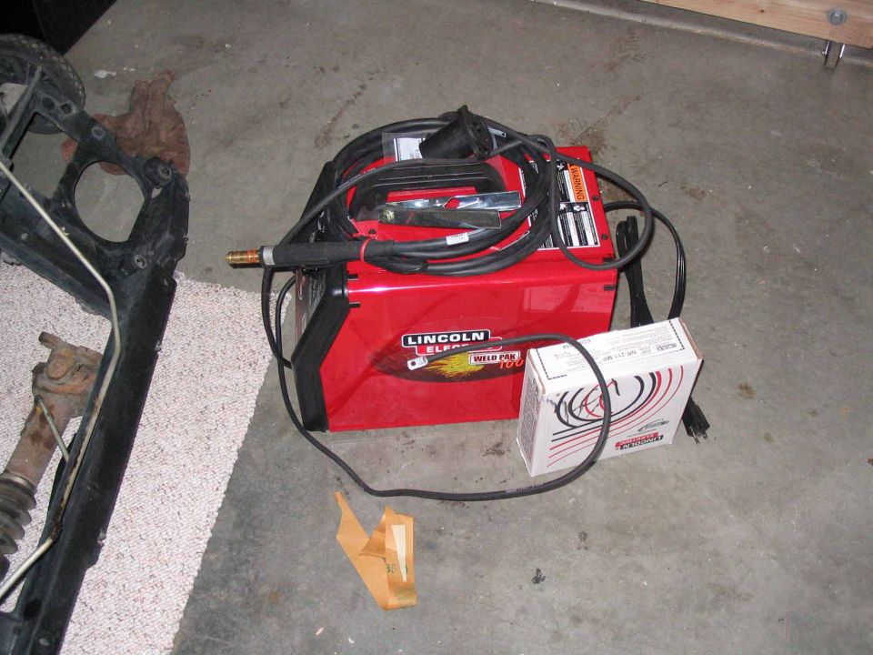 Red Lincoln Electric Weld Pak 100 welder with cables and a box of supplies on a concrete floor next to some vehicle parts