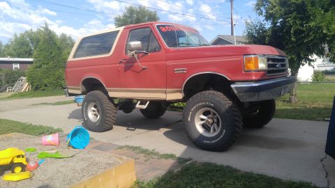 1991 Ford Bronco - This lifted Ford Bronco has large off-road tires and a tan stripe along its side.