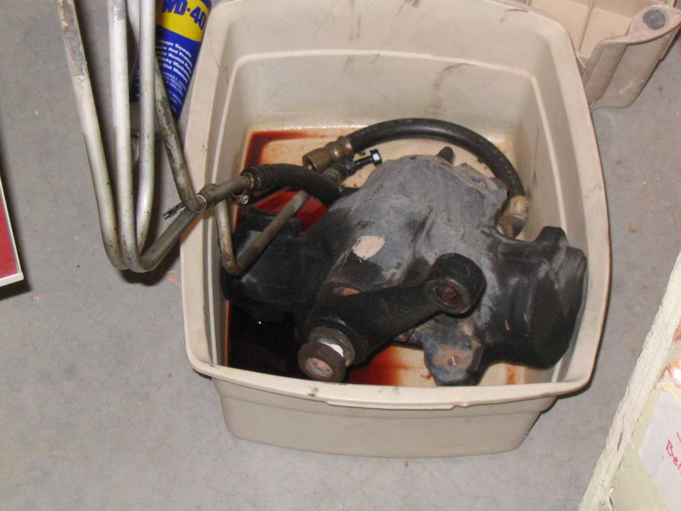 Power steering gearbox submerged in reddish fluid within a white plastic drain pan. Metal lines are attached