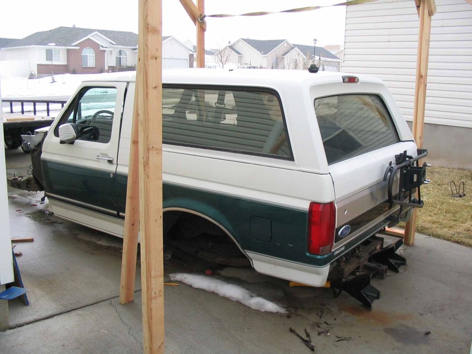 1996 Ford Bronco, white and green, undergoing repairs under a wooden frame. Rear-mounted spare tire visible
