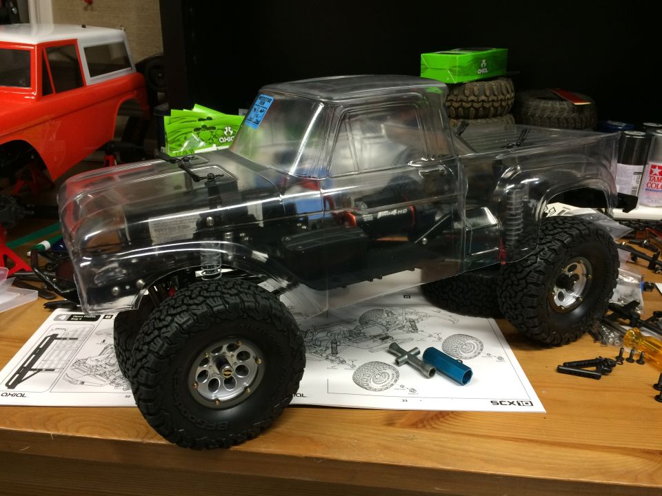 Clear plastic RC truck body with off-road tires on a workbench, next to an orange RC truck body