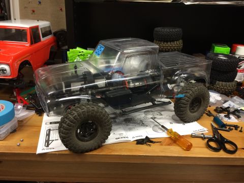 1989 Ford Bronco - An unassembled RC truck with a clear body sits on a workbench next to a red truck body, surrounded by tools and parts.