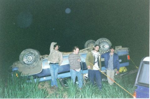 1981 Ford Bronco - Four people stand around a flipped blue truck at night, with one tire prominently displayed.