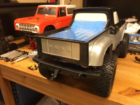 1989 Ford Bronco - Two RC car bodies, a silver and black pickup truck and an orange Bronco, sit on a wooden workbench.