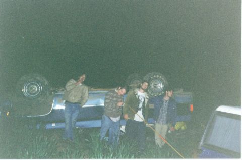 1981 Ford Bronco - A group of people stand around a vehicle that has flipped onto its side in a grassy area at night.