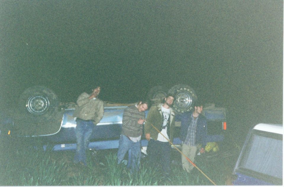 Blue truck upside down in a grassy field at night with four people standing beside it