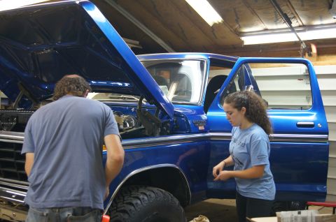 1979 Ford Bronco - Mechanics work on the engine of a blue 1979 Ford Bronco with its hood and driver's door open in a garage.