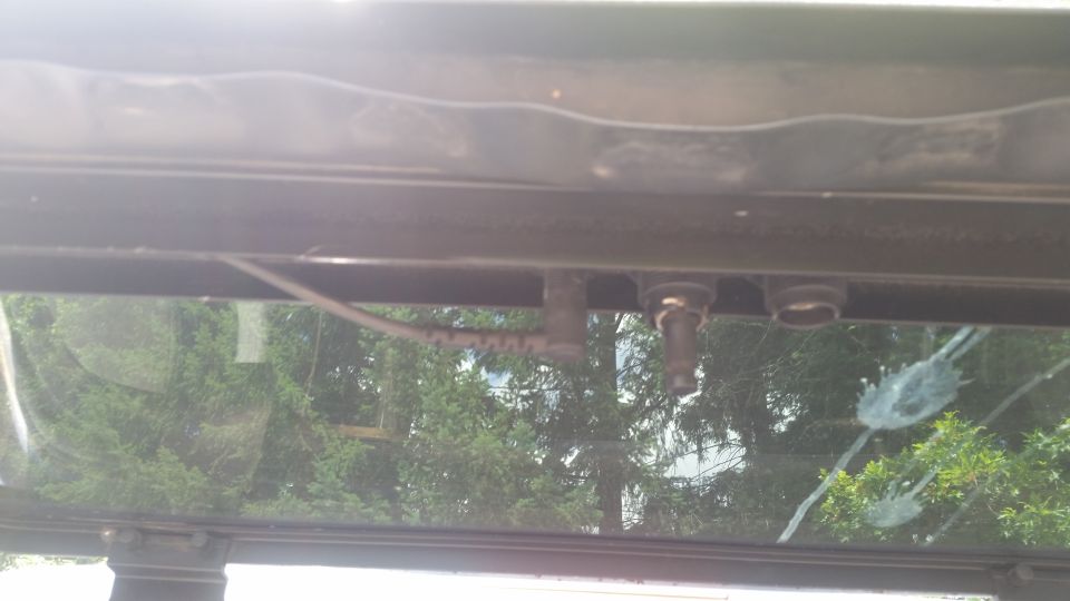 Underside of roof header above glass showing wiring and a small nozzle fitting, trees visible through window