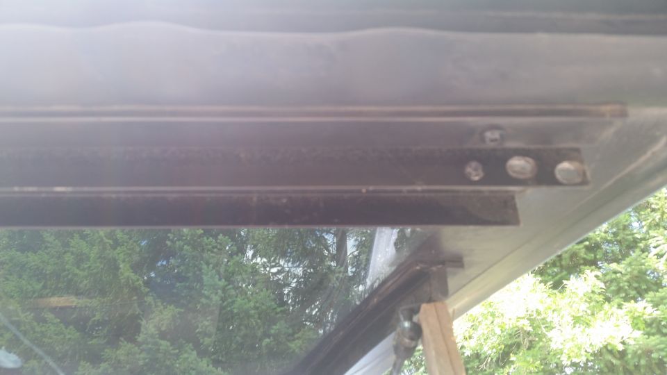 Underside of raised Bronco liftgate showing black bolted metal track, trees in background