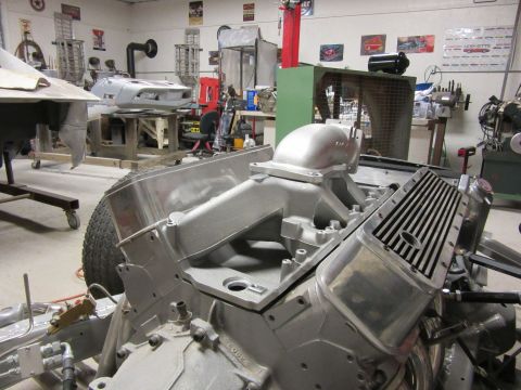Close-up of a V8 engine block and intake manifold, with a tire and chassis components visible. A car body is i