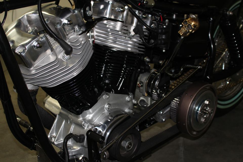 Close-up of a V-twin motorcycle engine with a black drive belt and a gold skull shift knob. Supermotors.net logo visible