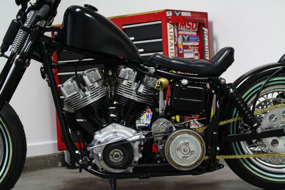 Black custom motorcycle with a V-twin engine and a skull shifter. The rear wheel has a gold chain and striped tire