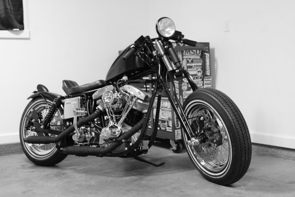 Custom motorcycle with black fuel tank and seat, chrome engine, and spoked wheels. Toolbox with stickers is in the background