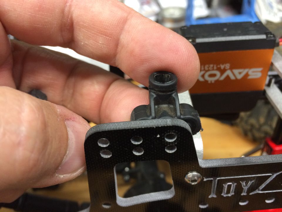 Hand holding a black plastic RC car suspension component with a metal screw. "TOYZ" is visible on the chassis