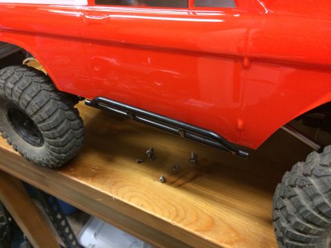1989 Ford Bronco - Side view of a red RC car body with installed rock sliders and scattered screws on a wooden workbench.