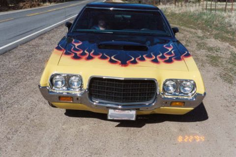 1973 Ford Ranchero - A yellow car with flame decals on the hood is parked on a dirt road next to a paved road.