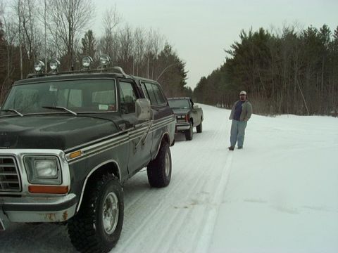 1978 Ford Bronco - Two vehicles drive down a snowy road in a wooded area, with a man standing beside the road.