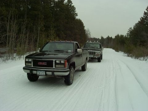 1978 Ford Bronco - Two pickup trucks drive down a snowy road surrounded by trees.