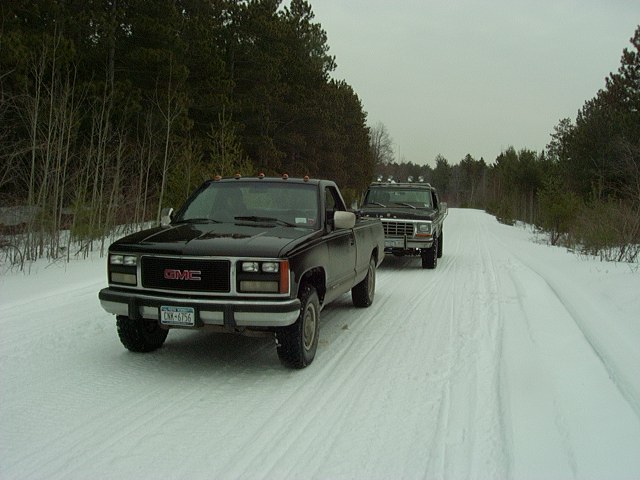 Two pickup trucks on a snow-covered road, one black GMC in the foreground, a darker truck behind it