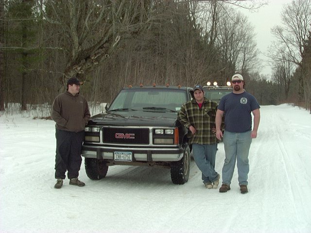 Three men stand by two GMC trucks in a snowy, wooded area. The front truck is black with a GMC grille and a license plate reading CMX-6756
