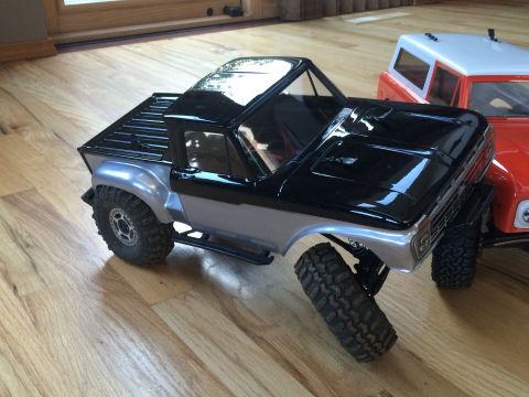1989 Ford Bronco - Two remote-controlled trucks are parked on a wooden floor, one in the foreground and another partially visible behind it.
