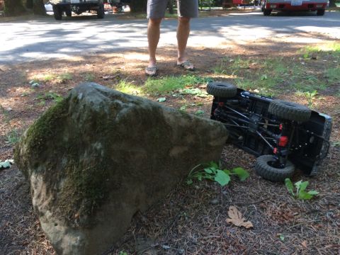 1989 Ford Bronco - An overturned remote control rock crawler lies on the ground near a large, moss-covered boulder.