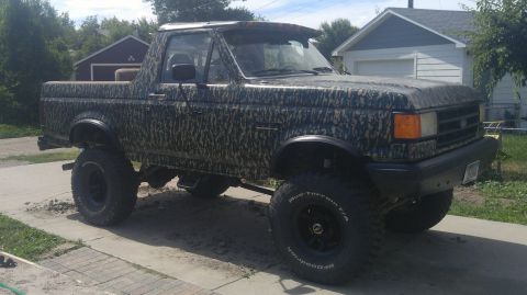 1991 Ford Bronco - A lifted Ford Bronco with camouflage wrap sits on a paved surface in front of a residential garage.