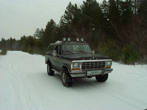 1978 Ford Bronco - A black 1978 Ford Bronco equipped with roof lights navigates a snow-covered road through a wooded area.