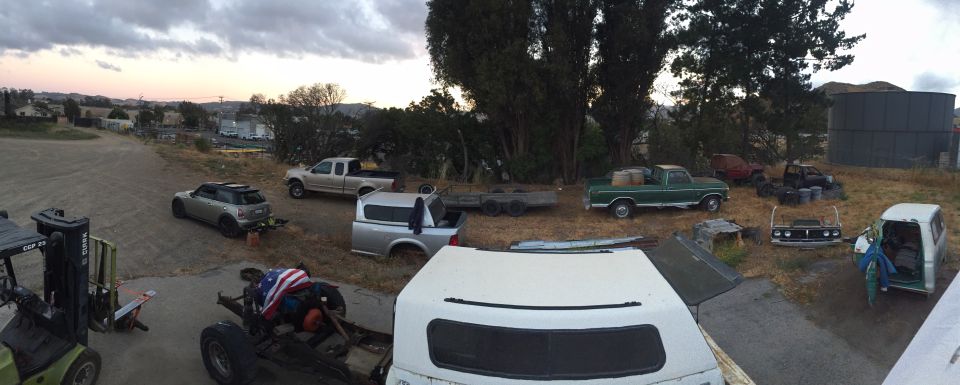 Panoramic view of a junkyard with various vehicles and parts, including a forklift and truck cap.