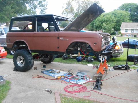 1972 Ford Bronco - A rusty brown 1972 Ford Bronco is undergoing repairs outdoors. The vehicle is lifted on jack stands, with the hood open and various tools a…