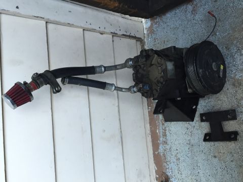 1989 Ford Bronco - Photograph of an automotive AC compressor with mounting bracket and a small cone filter attached, sitting on the ground.