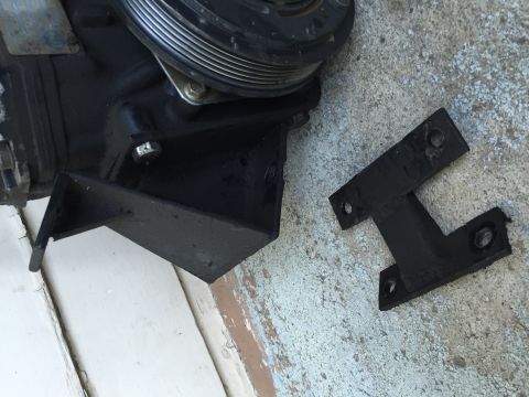 1989 Ford Bronco - Photograph of an AC compressor mounting bracket and a small H-shaped metal mounting plate on a concrete floor.