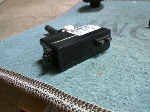 2007 Ford Crown Victoria - A black electronic module with a white label is shown on a teal carpeted surface. The module has two visible buttons labeled 'H' and 'M'.