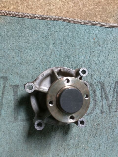 2007 Ford Crown Victoria - A metal automotive water pump with a black rubber seal is shown from a top-down angle. The part has several bolt holes and visible text inc…