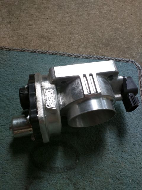 2007 Ford Crown Victoria - A silver throttle body with a black electrical connector is shown from a slightly elevated side angle. The part is resting on a textured te…