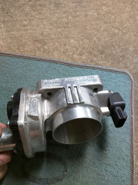 2007 Ford Crown Victoria - A silver BBK Power-Plus 70mm throttle body is shown from a three-quarter front angle, resting on a teal mat with a textured concrete backgr…