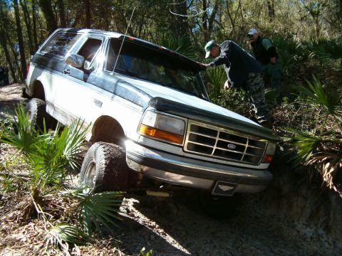 1991 Ford Bronco - A Ford Bronco navigates a muddy, wooded trail with two people observing.