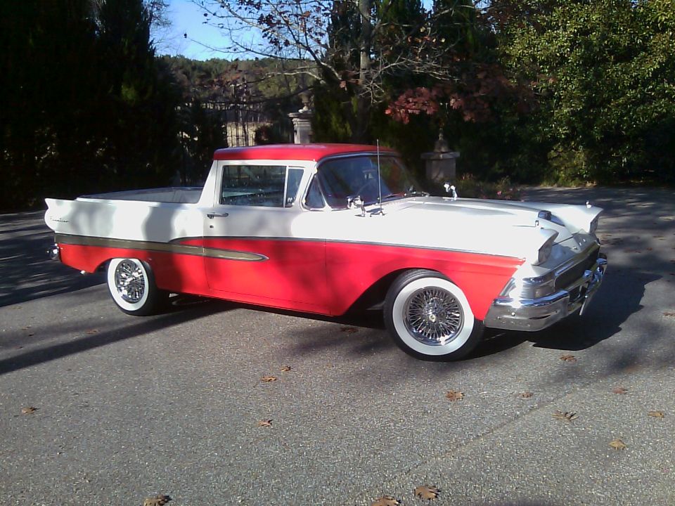 Red and white 1958 Ford Ranchero parked on a paved surface with trees in the background